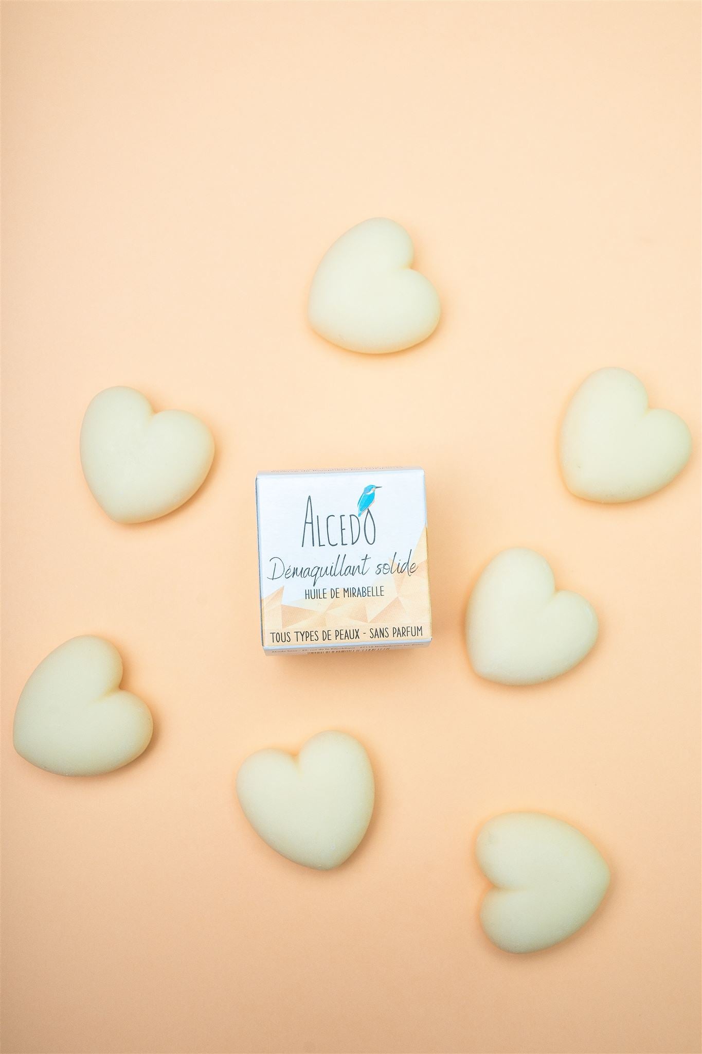 Boîte de démaquillant solide Alcedo à l’huile de mirabelle, entourée de démaquillant bio Alcedo en forme de cœur sur fond beige.