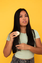 Femme aux cheveux longs tenant un après-shampoing Alcedo dans la main, sur fond jaune uni.