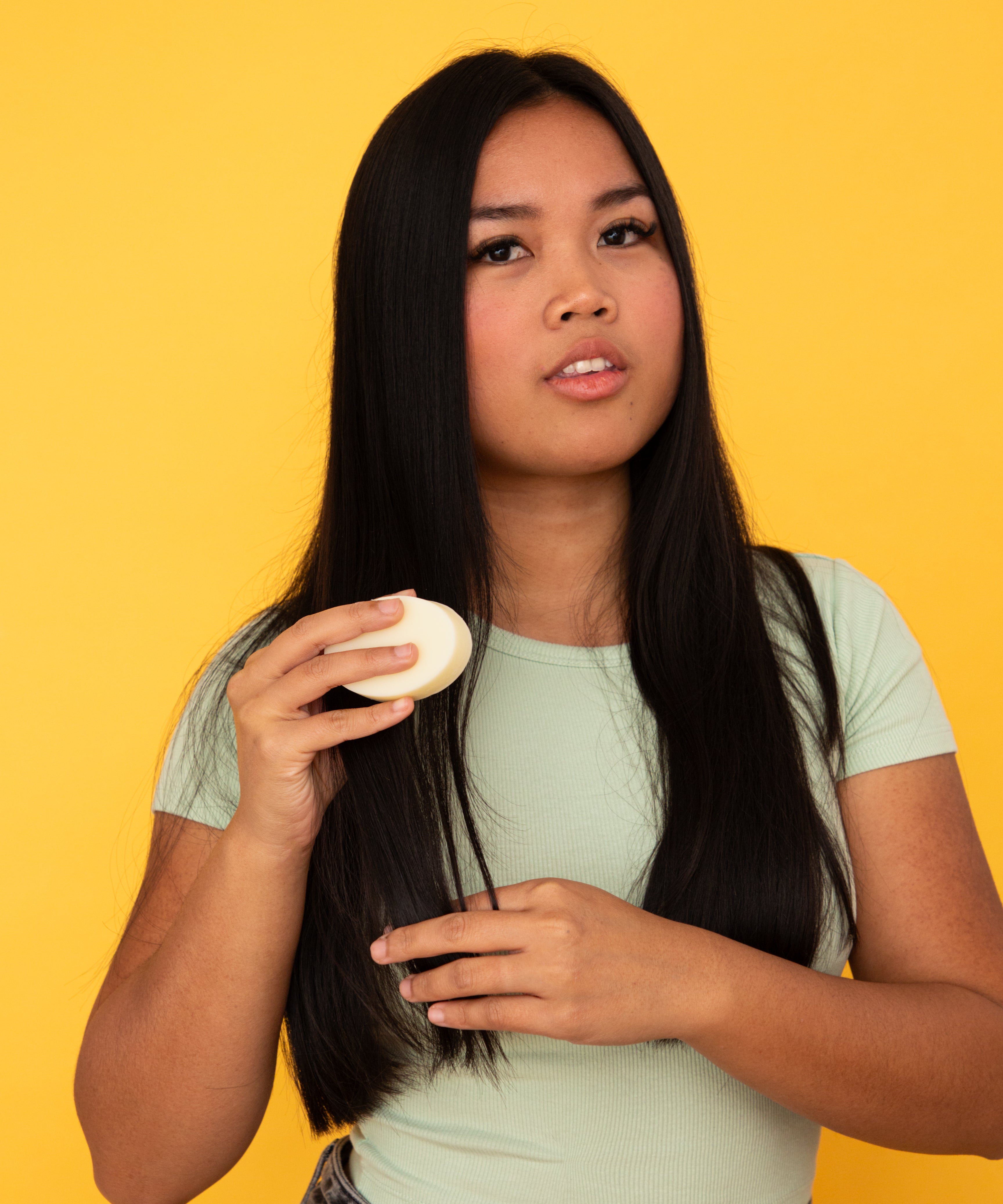 Femme aux cheveux longs tenant un après-shampoing Alcedo dans la main, sur fond jaune uni.