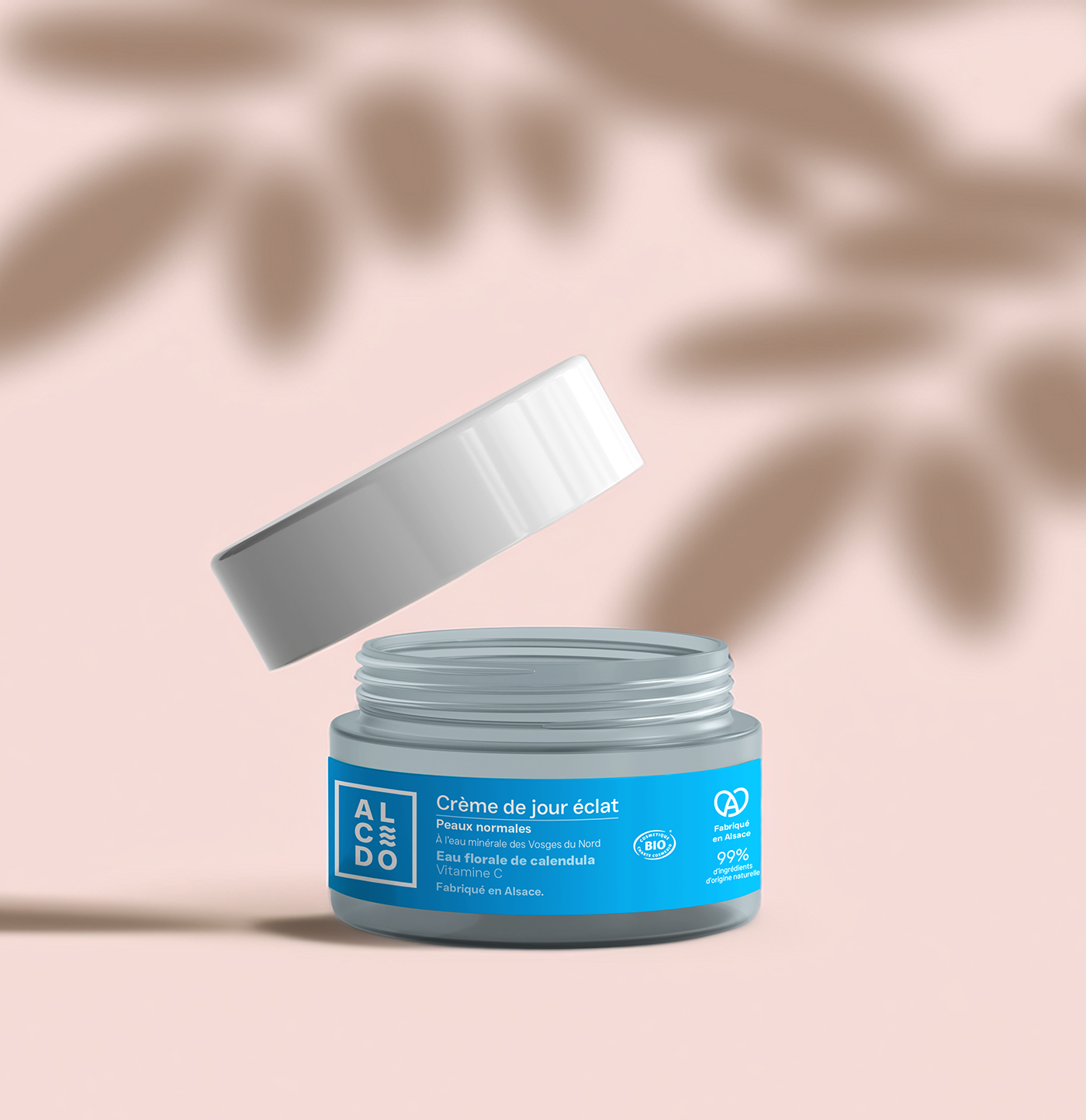 Pot de crème de jour éclat Alcedo ouvert avec le couvercle légèrement soulevé, sur fond beige.