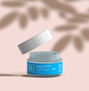 Pot de crème de jour éclat Alcedo ouvert avec le couvercle légèrement soulevé, sur fond beige.