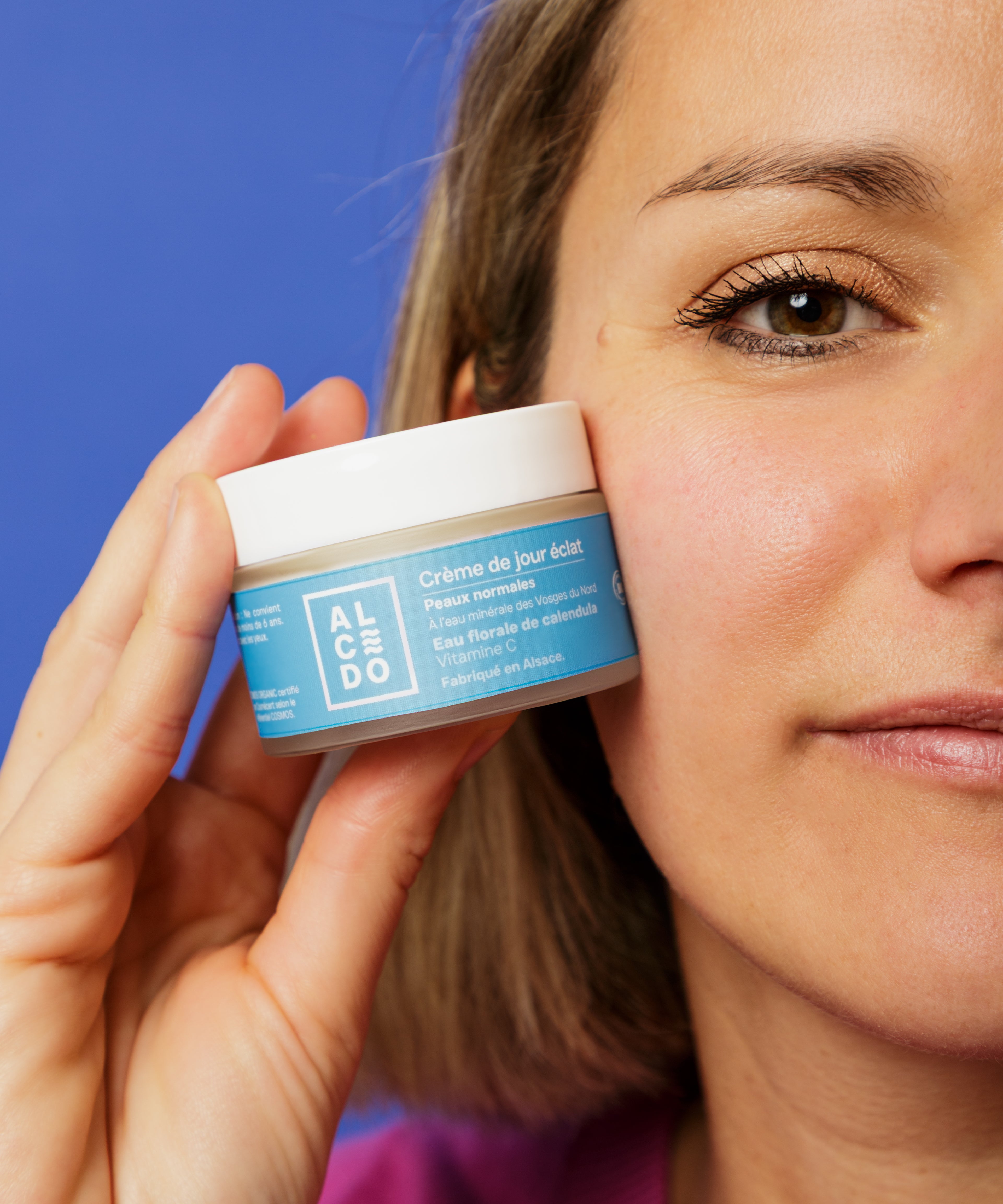 Gros plan sur Maud Steininger - fondatrice de la marque Alcedo, tenant un pot de crème de jour éclat Alcedo contre sa joue, avec un fond bleu uni.