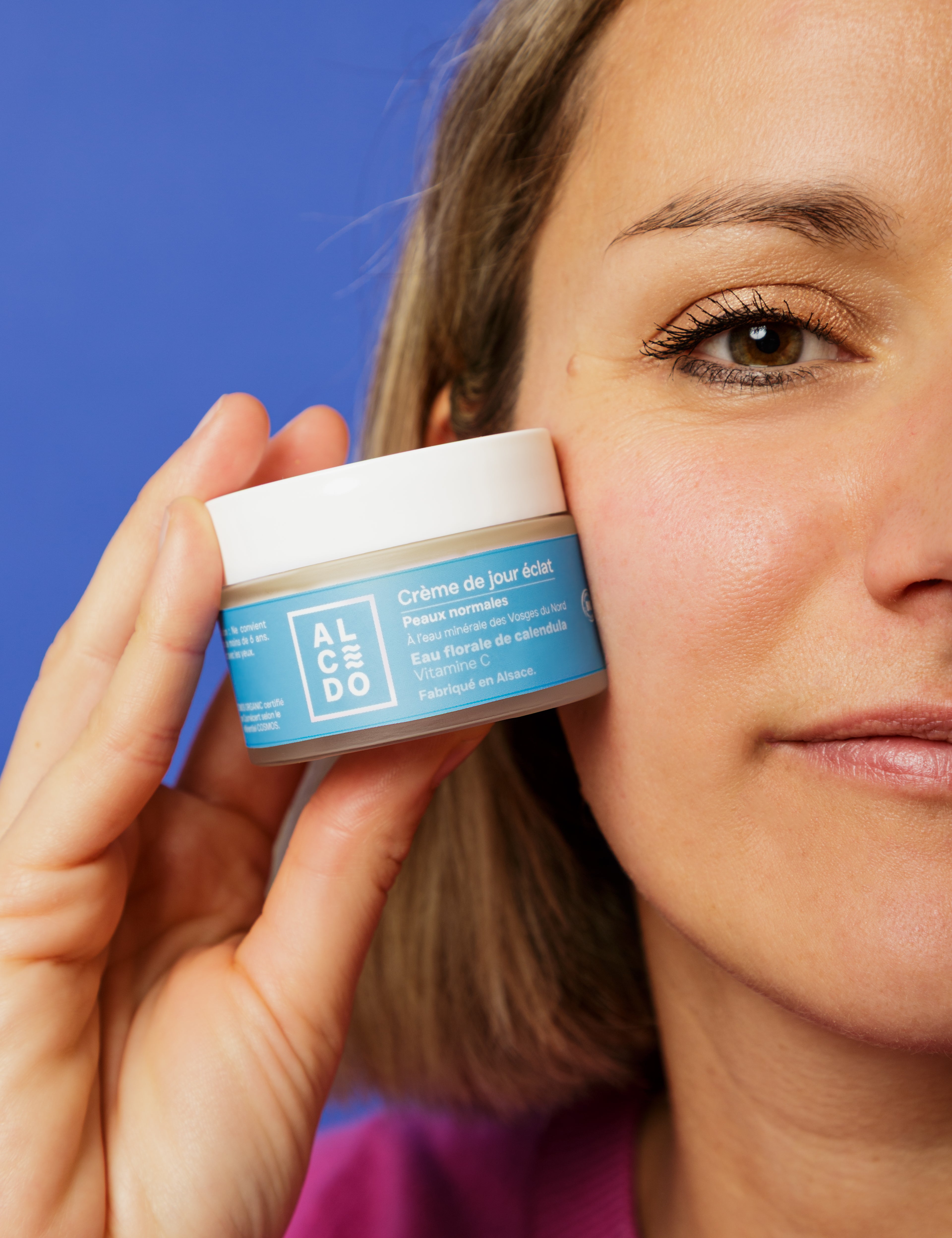 Gros plan sur Maud Steininger - fondatrice de la marque Alcedo, tenant un pot de crème de jour éclat Alcedo contre sa joue, avec un fond bleu uni.