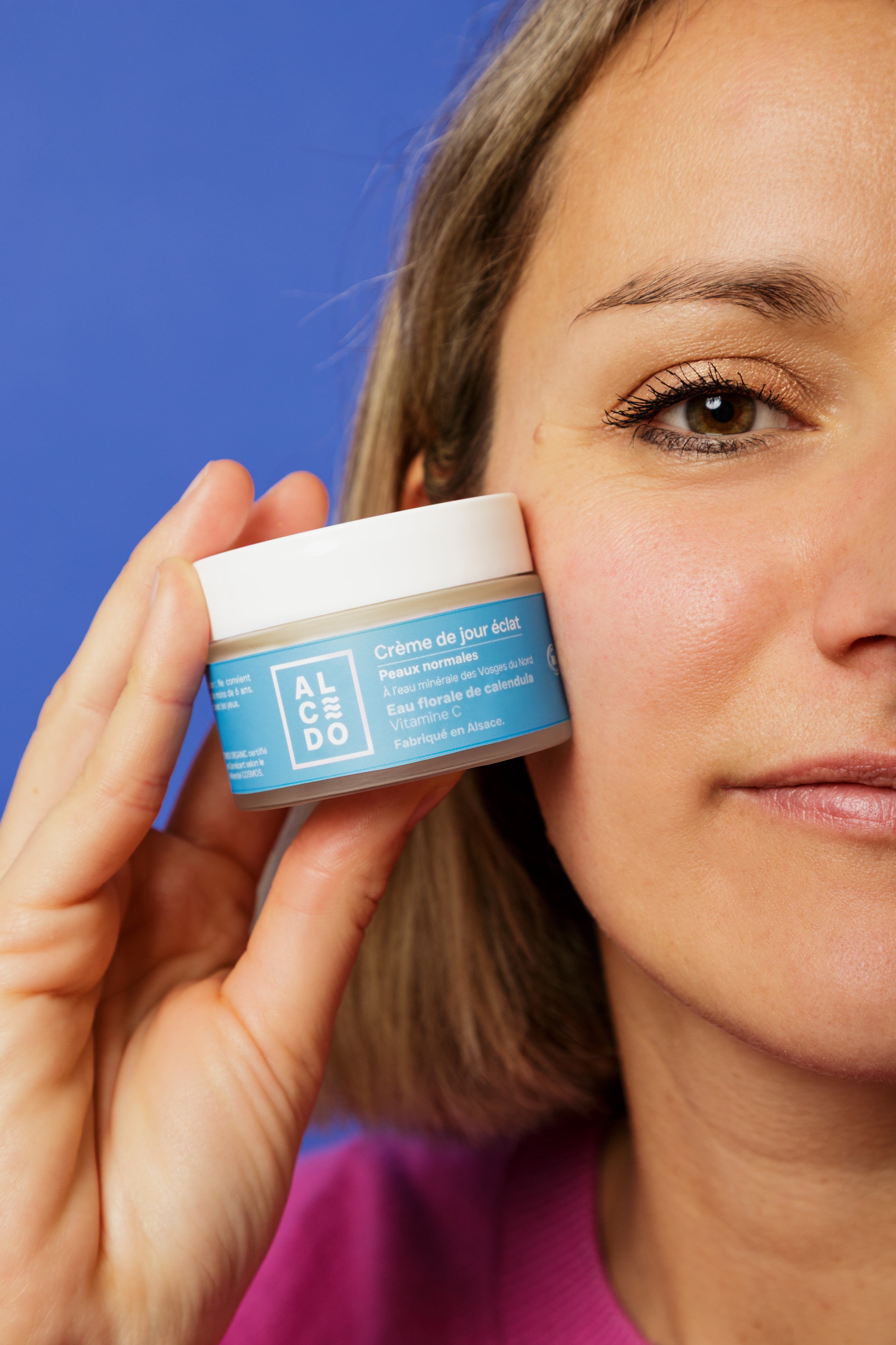 Gros plan sur Maud Steininger - fondatrice de la marque Alcedo, tenant un pot de crème de jour éclat Alcedo contre sa joue, avec un fond bleu uni.