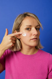 Gros plan sur Maud Steininger (la fondatrice de la marque Alcedo) appliquant une noisette de crème sur sa joue avec le doigt, devant un fond bleu uni.