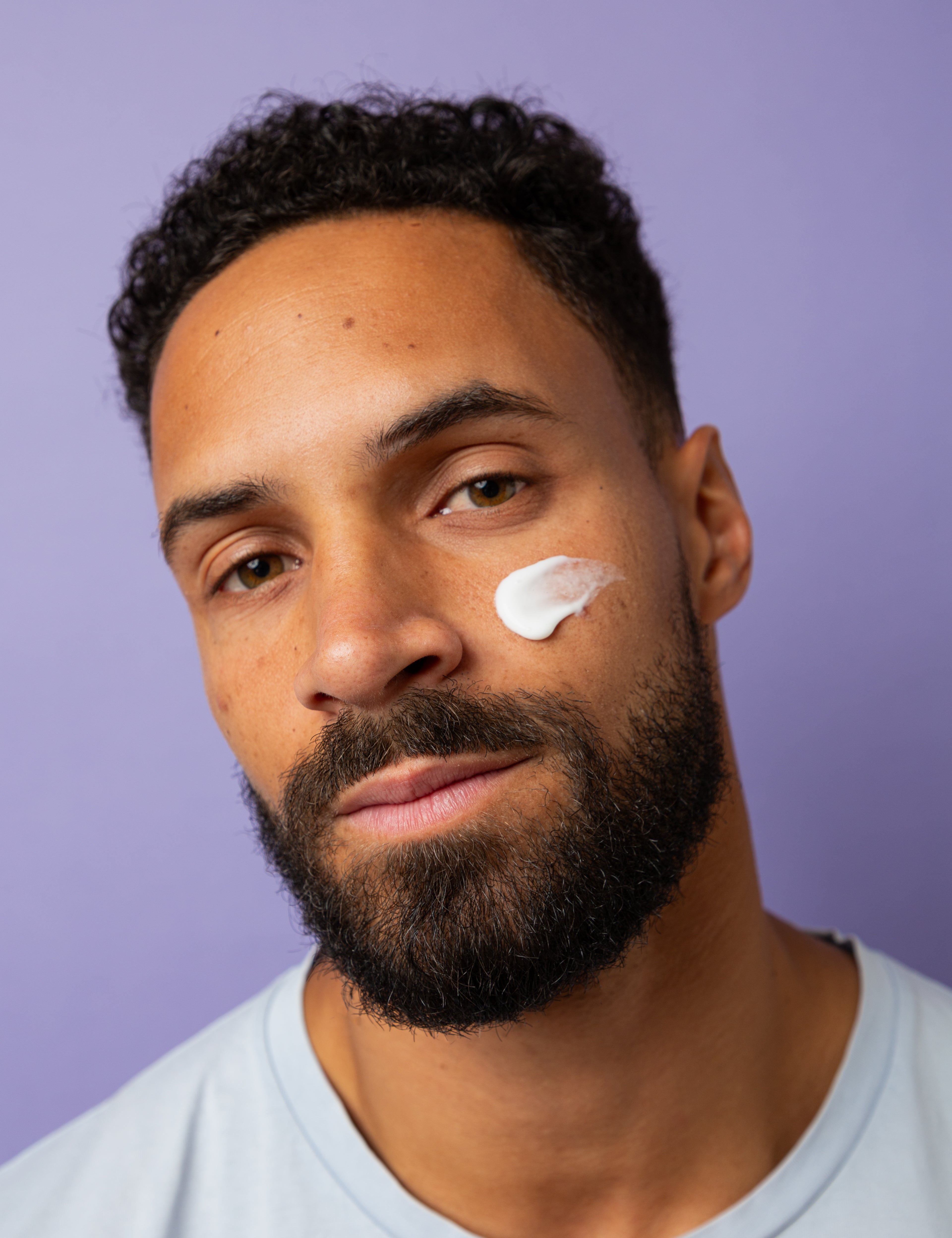 Gros plan sur un homme portant une noisette de crème hydratante de Alcedo sur la joue, regard face caméra, sur fond violet clair.