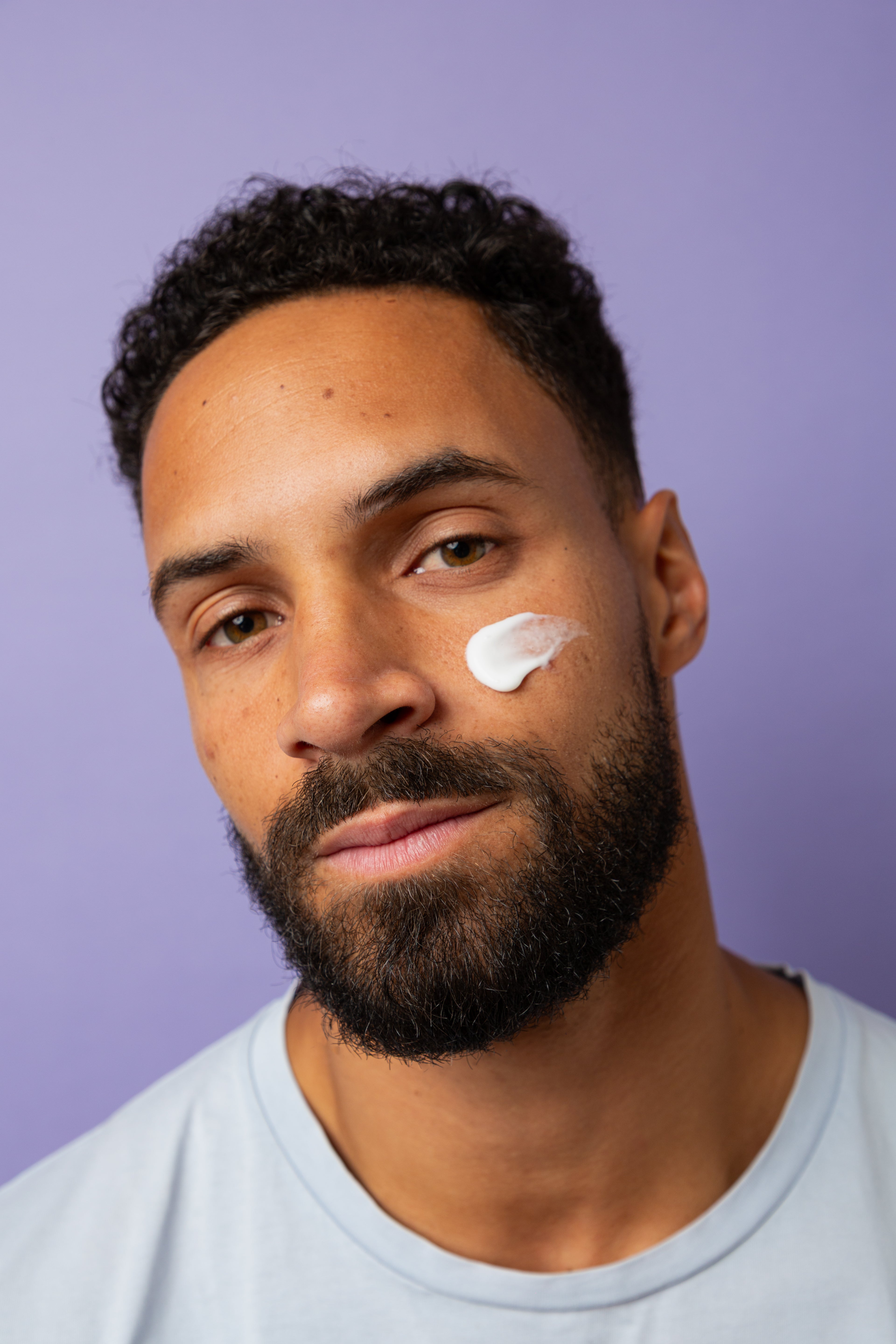 Gros plan sur un homme portant une noisette de crème hydratante de Alcedo sur la joue, regard face caméra, sur fond violet clair.