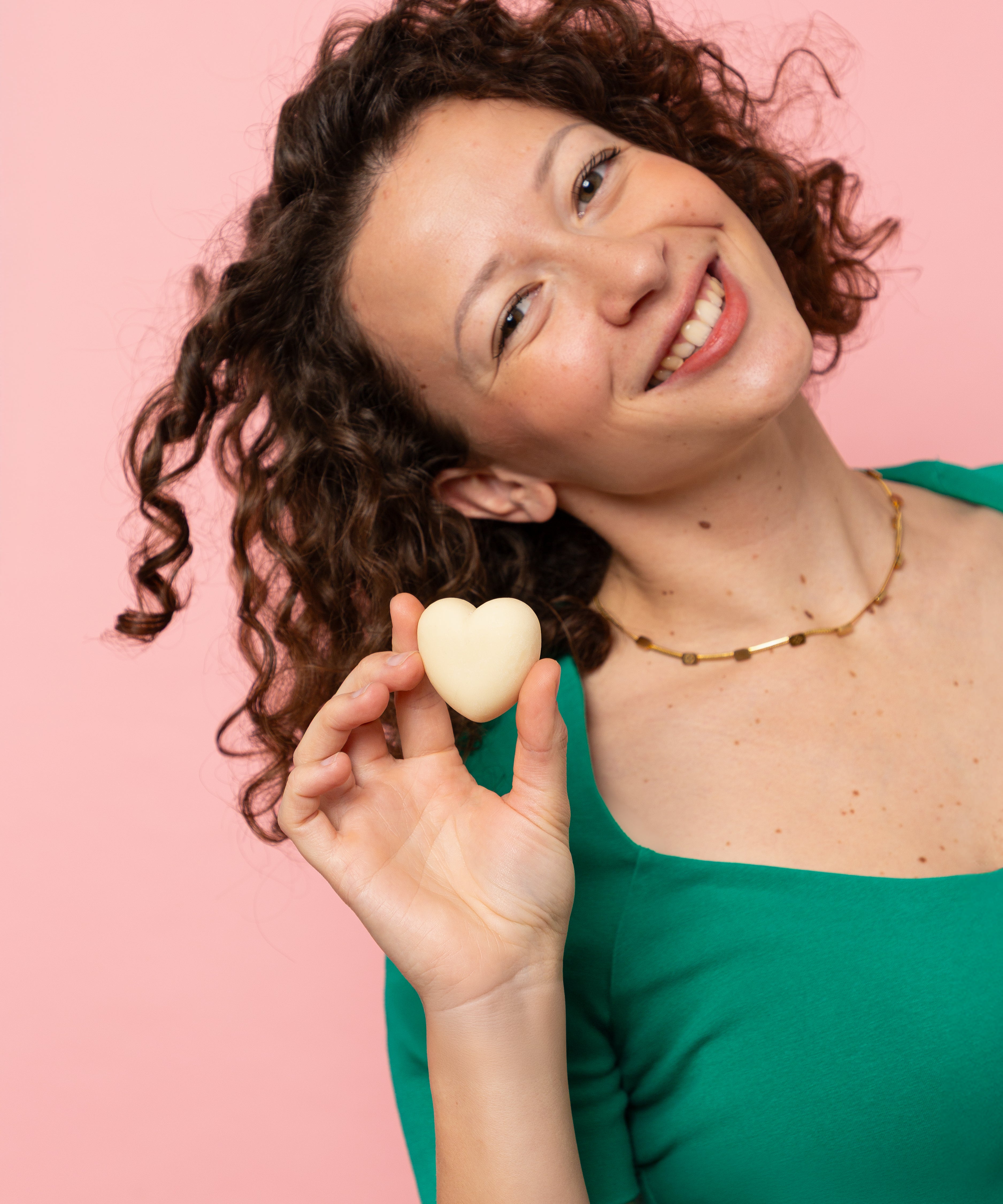 Femme souriante aux cheveux bouclés tenant un démaquillant solide Alcedo en forme de cœur devant un fond rose