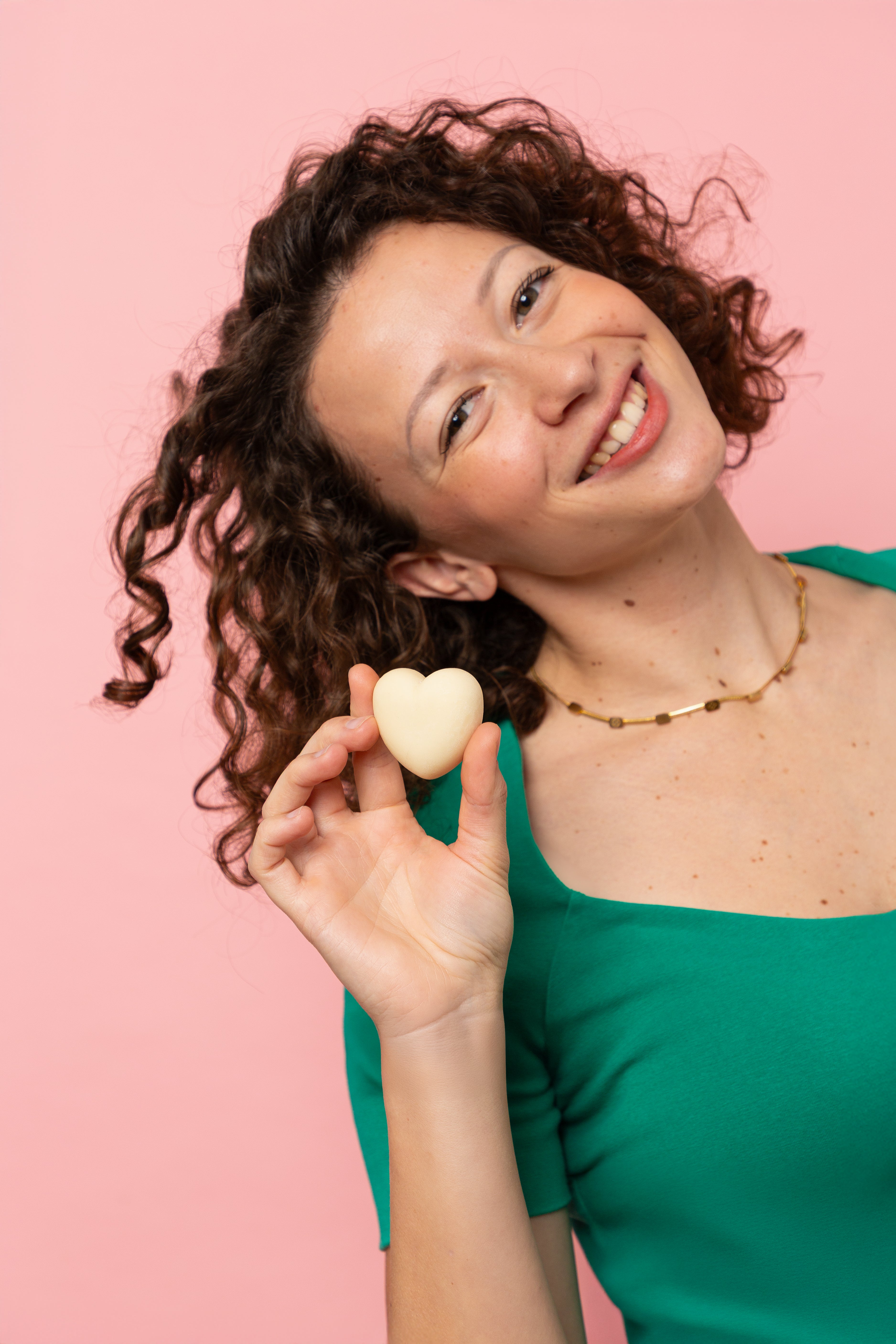 Femme souriante aux cheveux bouclés tenant un démaquillant solide Alcedo en forme de cœur devant un fond rose