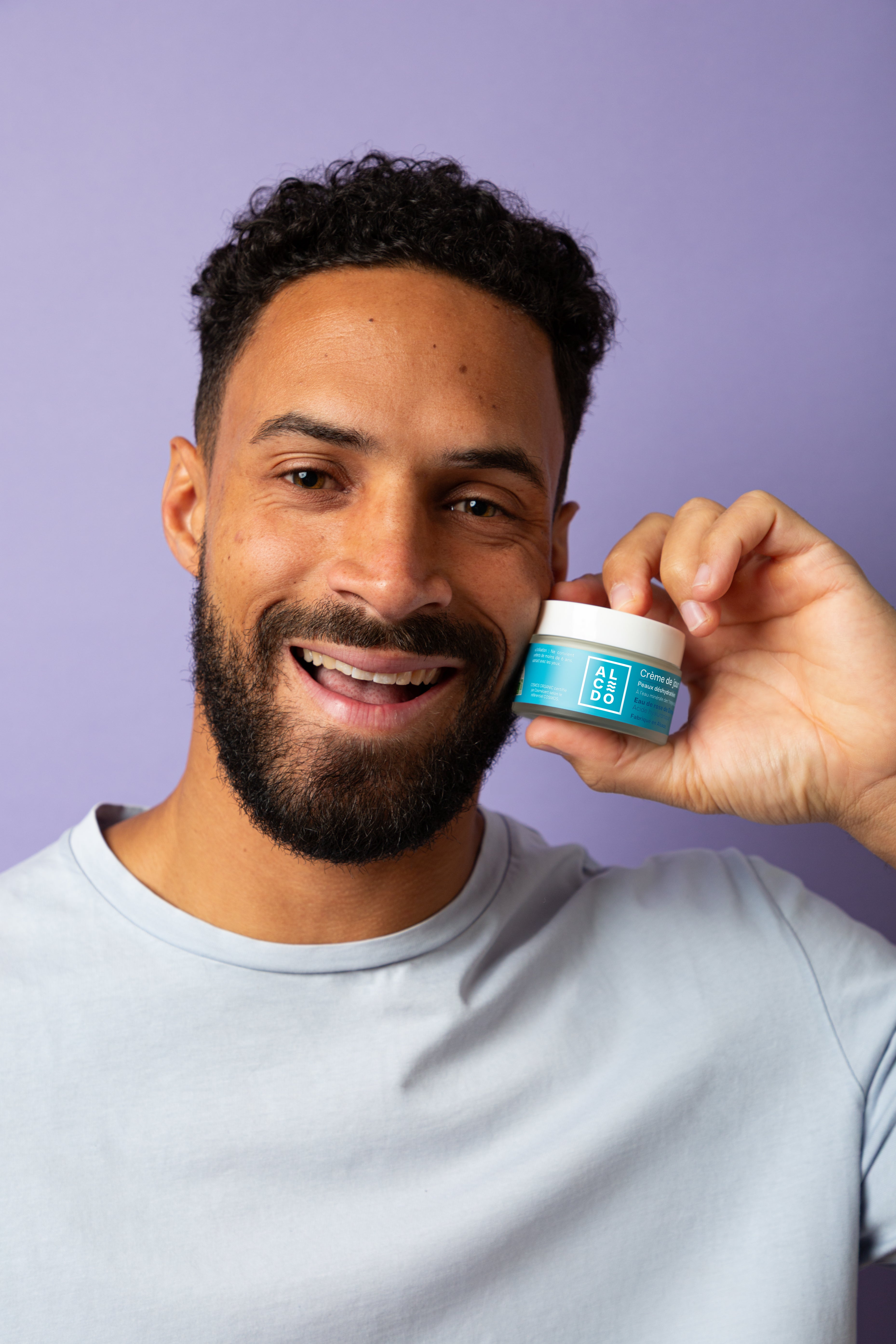 Homme souriant tenant un pot de crème Alcedo près de son visage, sur fond violet.